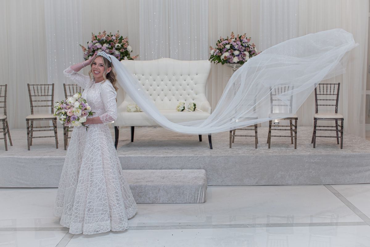 A smiling bride adjusts her billowing veil on a wedding stage, holding a bouquet near floral arrangements and wooden chairs.