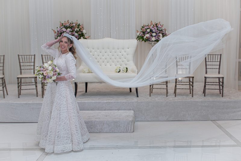 A smiling bride adjusts her billowing veil on a wedding stage, holding a bouquet near floral arrangements and wooden chairs.