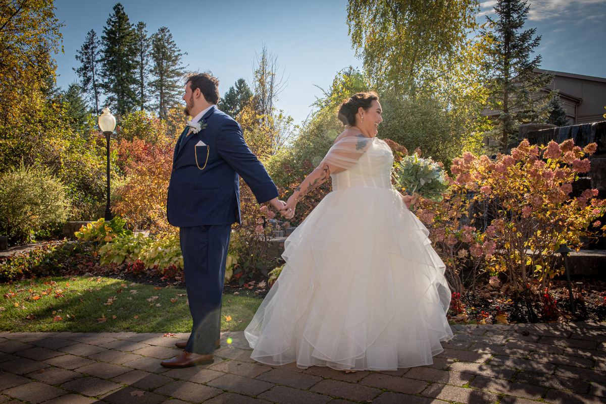 A newlywed couple in autumn walks along a brick path, the bride in a white gown with tattoos visible.
