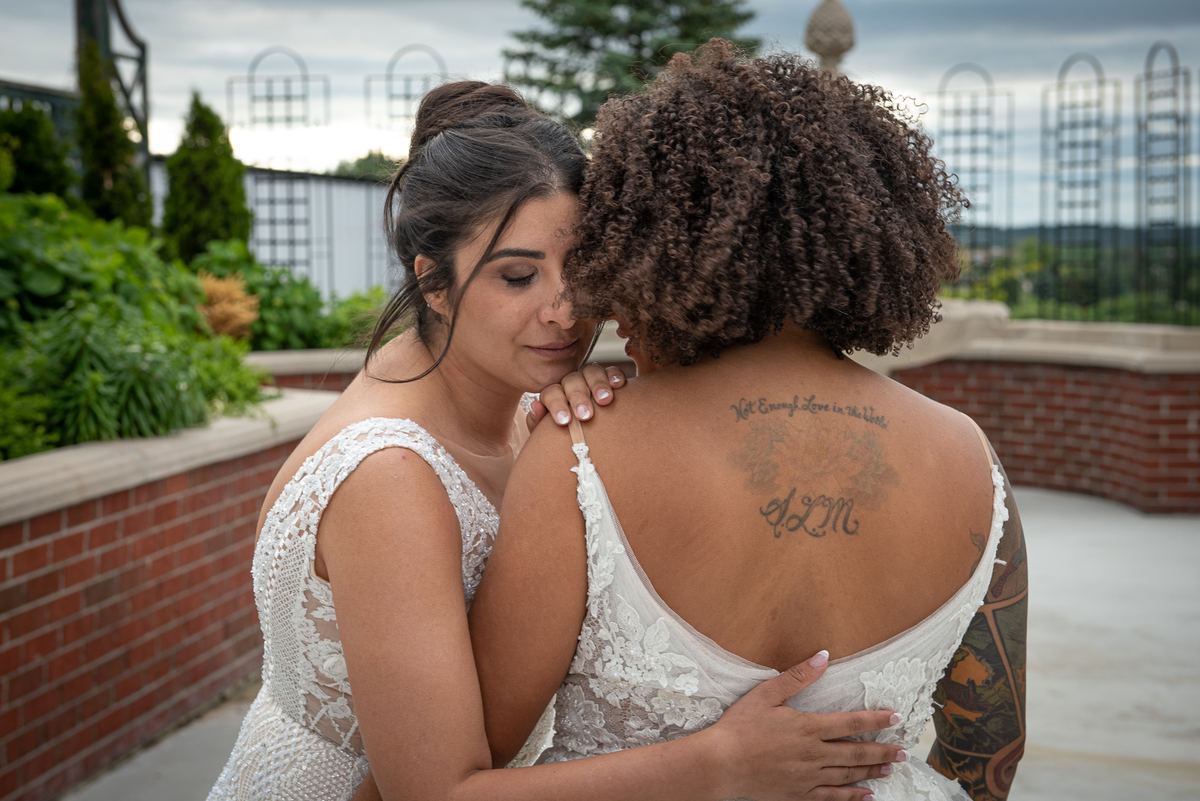 Two brides embrace intimately outdoors, one resting her head on the other's shoulder, illuminated by soft daylight.