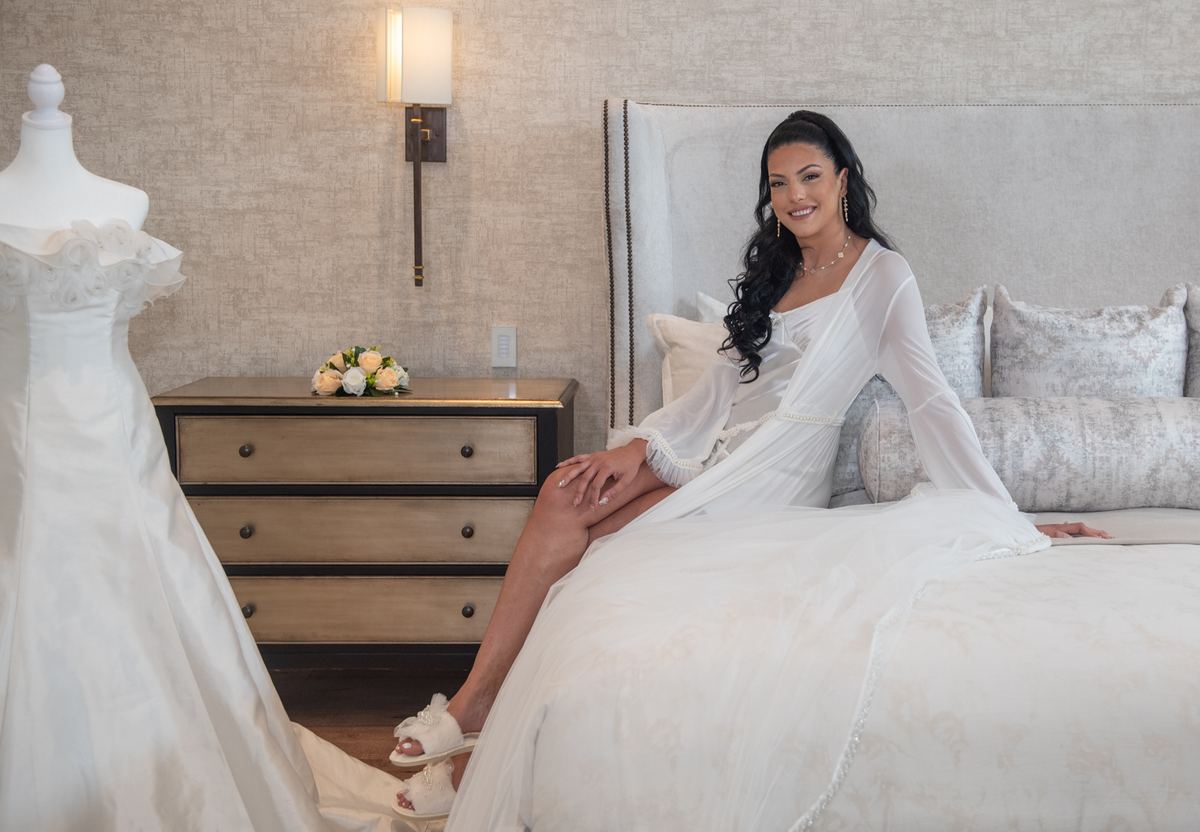 A bride smiles warmly while seated on a bed in an elegant bedroom, wearing a flowing white robe and sandals.