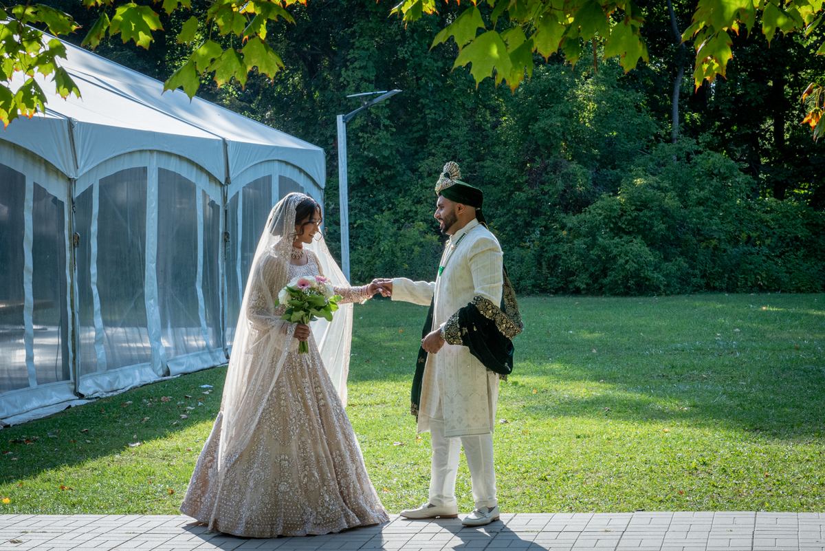 A bride in a lehenga and veil stands with her groom in a sherwani under dappled sunlight, holding hands.
