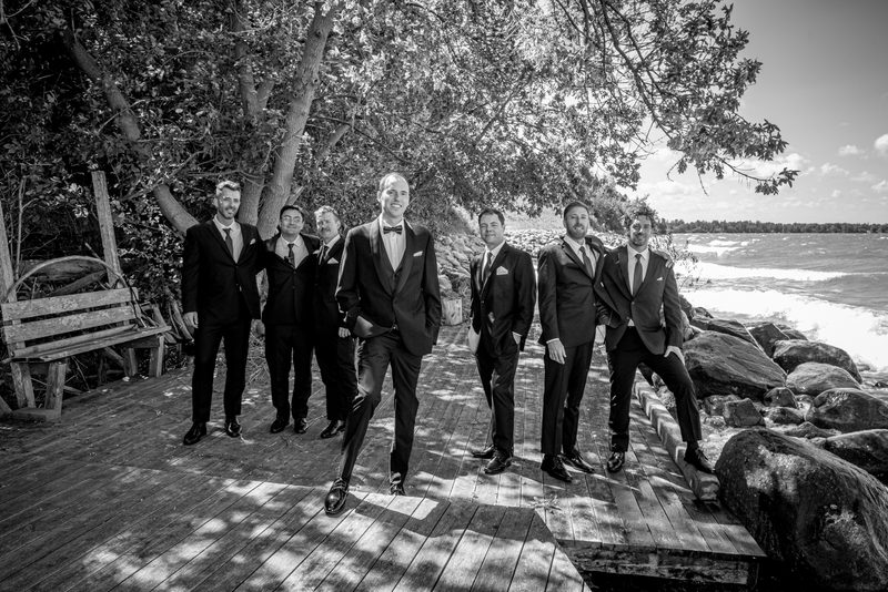 A groom and his groomsmen pose confidently on a wooden dock beside rocks, with sunlight filtering through trees.