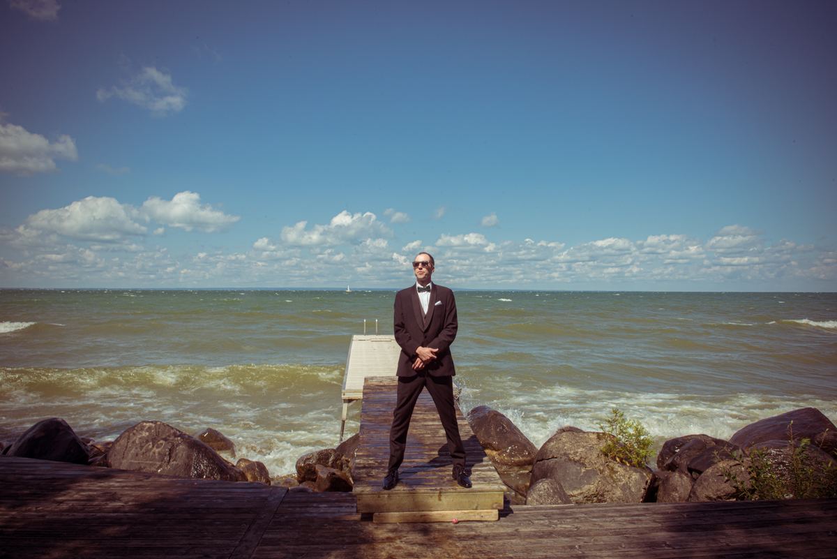 A man in a tuxedo with sunglasses stands on a wooden pier by a choppy sea under a partly cloudy sky.