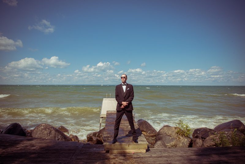 A man in a tuxedo with sunglasses stands on a wooden pier by a choppy sea under a partly cloudy sky.