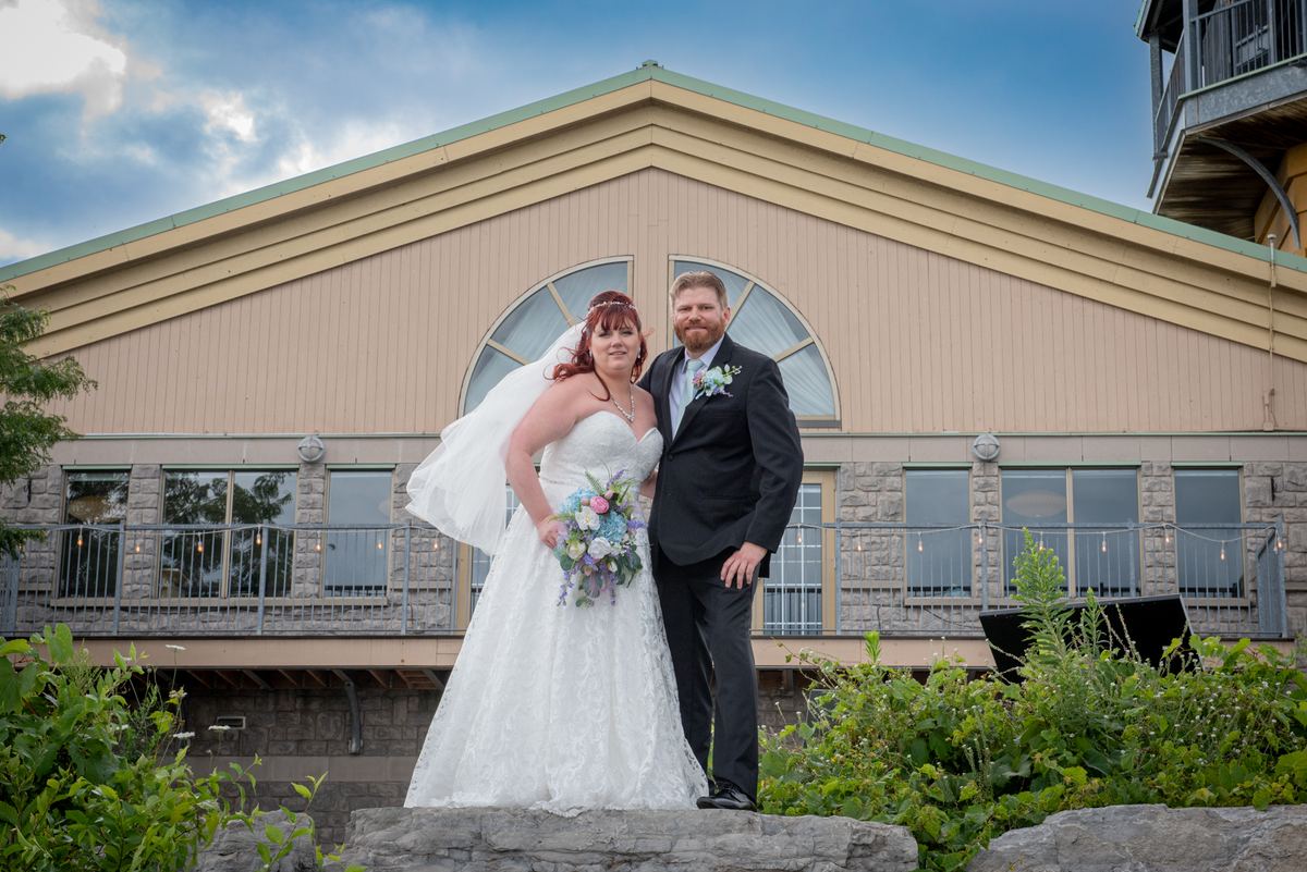 A bride and groom stand on a stone ledge in front of a modern building, surrounded by lush greenery.