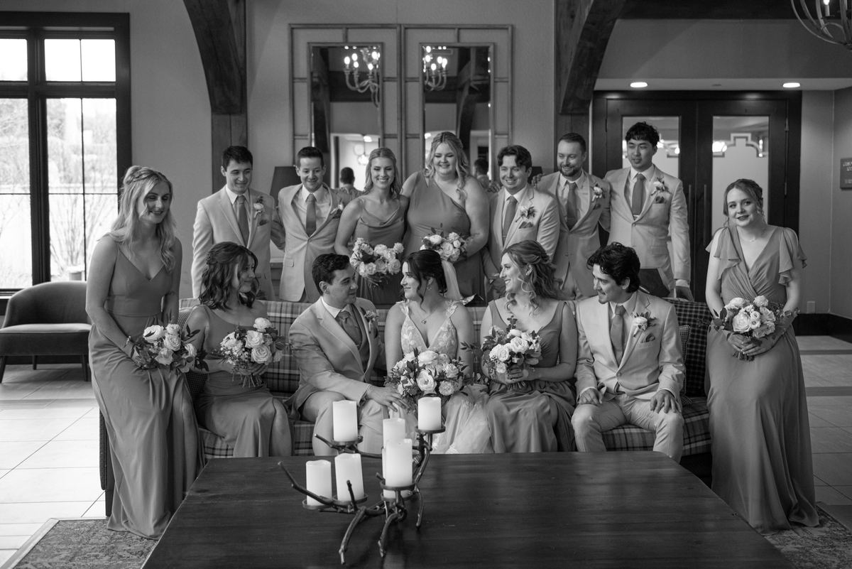 A joyful wedding party in an elegant indoor space, with the bride and groom surrounded by smiling attendants.