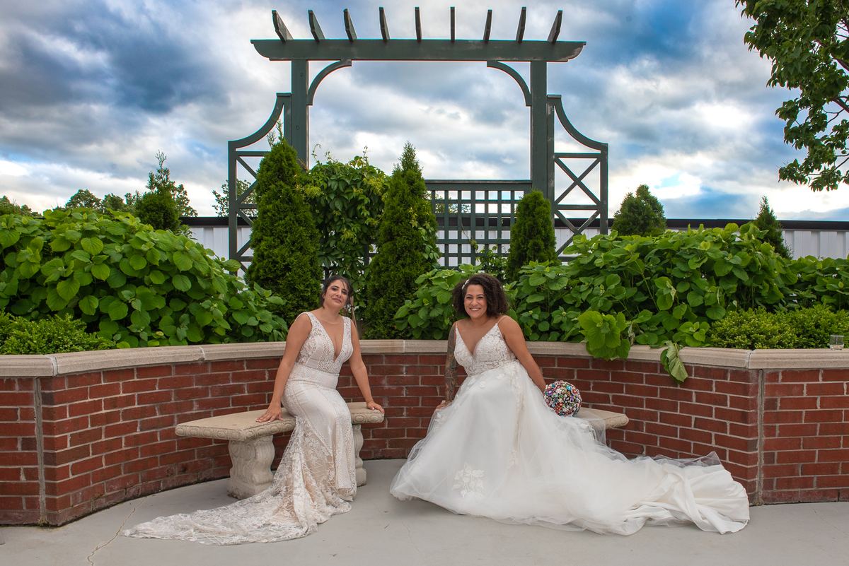 Two brides in elegant white gowns and lace smile beside a brick wall in a lush garden under a cloudy sky.