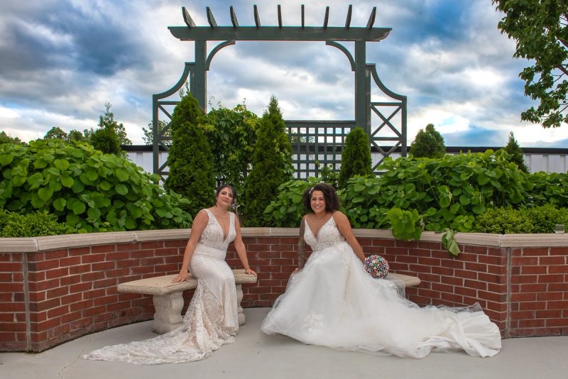 Two brides in elegant white gowns and lace smile beside a brick wall in a lush garden under a cloudy sky.