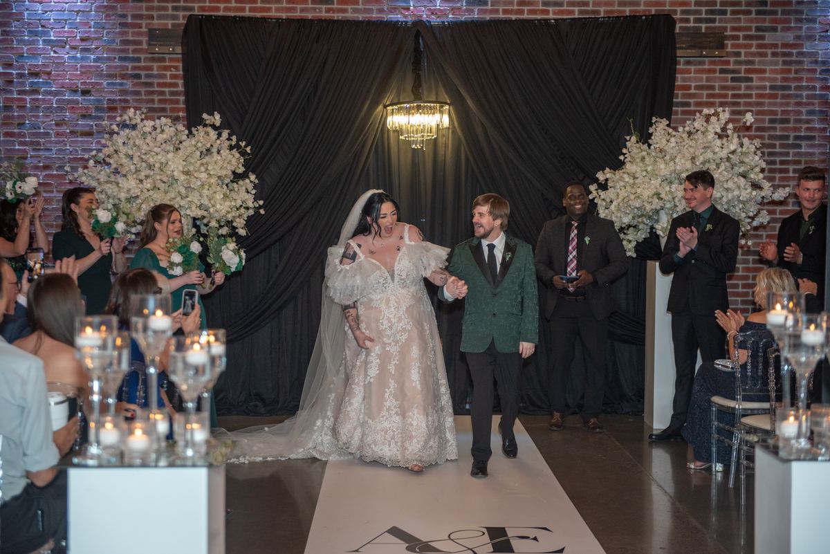 A smiling bride and groom walk down the aisle, surrounded by guests and floral arrangements in warm lighting.