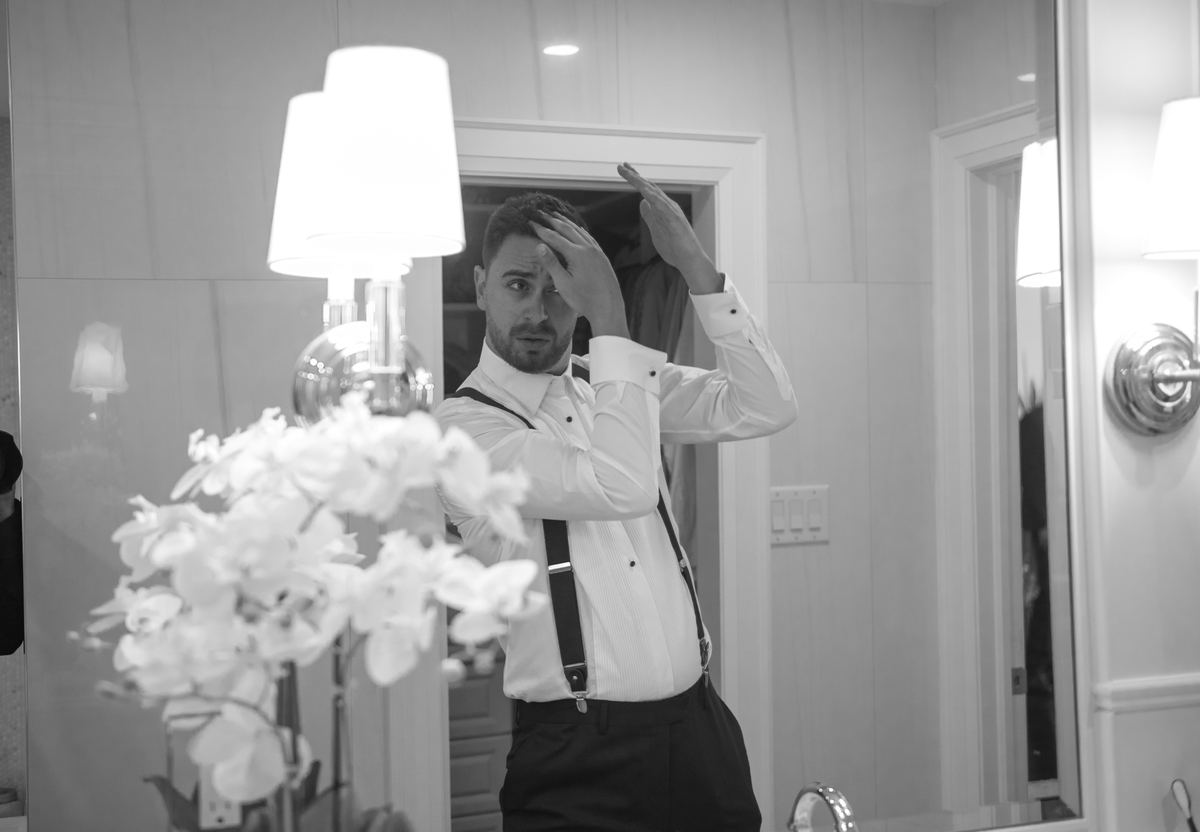 A man stands before a mirror in a well-lit room, adjusting his hair with one hand while looking intently at his reflection.