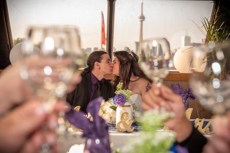 A newlywed couple kisses at their reception table adorned with purple and white flowers, as guests toast in the background.