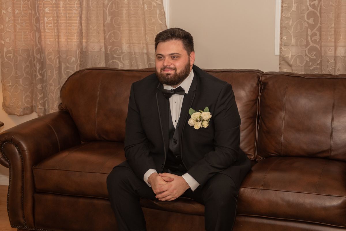 A groom in a black tuxedo with a boutonnière sits on a leather sofa, smiling warmly near patterned curtains.