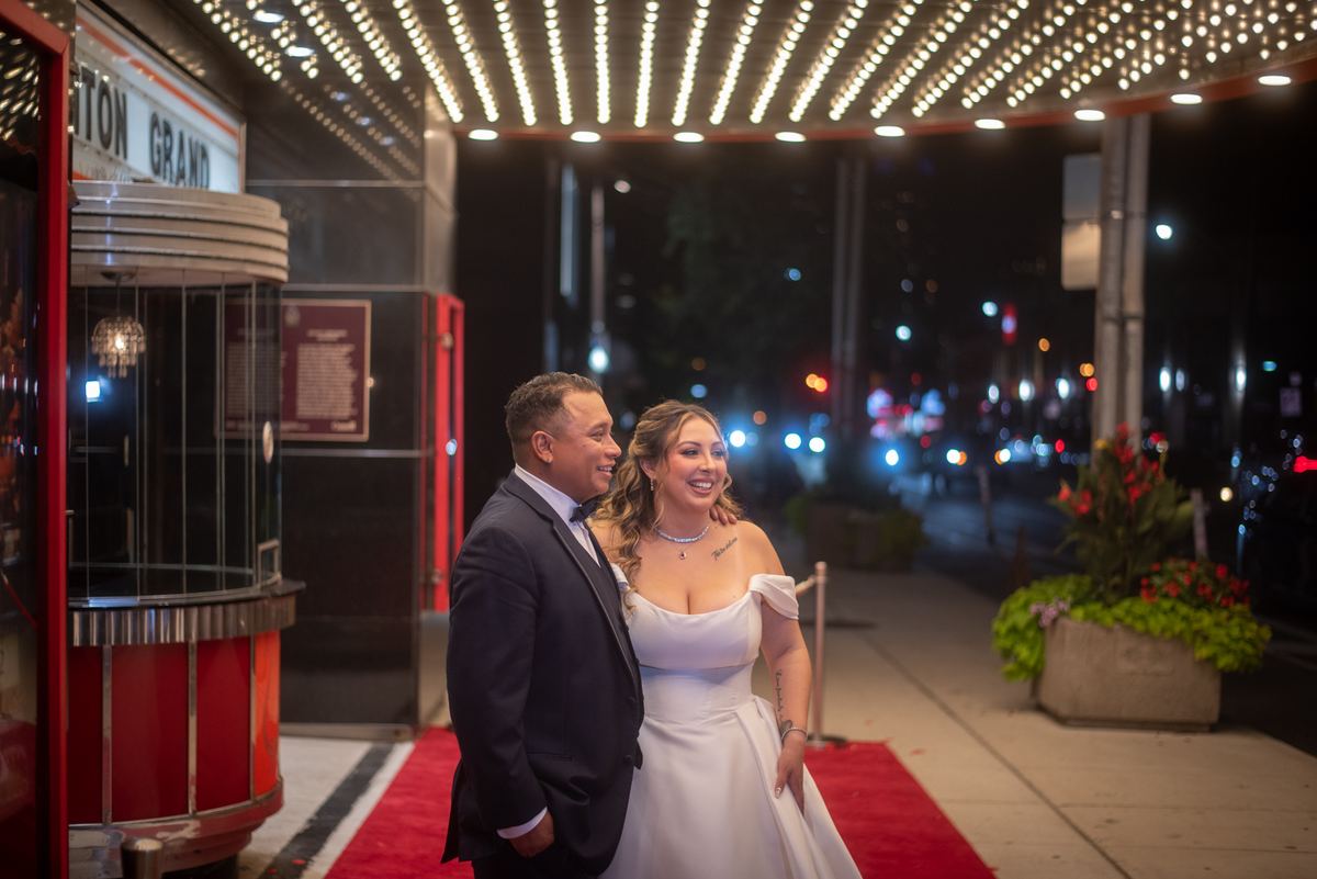 A newlywed couple stands on a red carpet outside a theater at night, smiling warmly under the bright marquee lights.