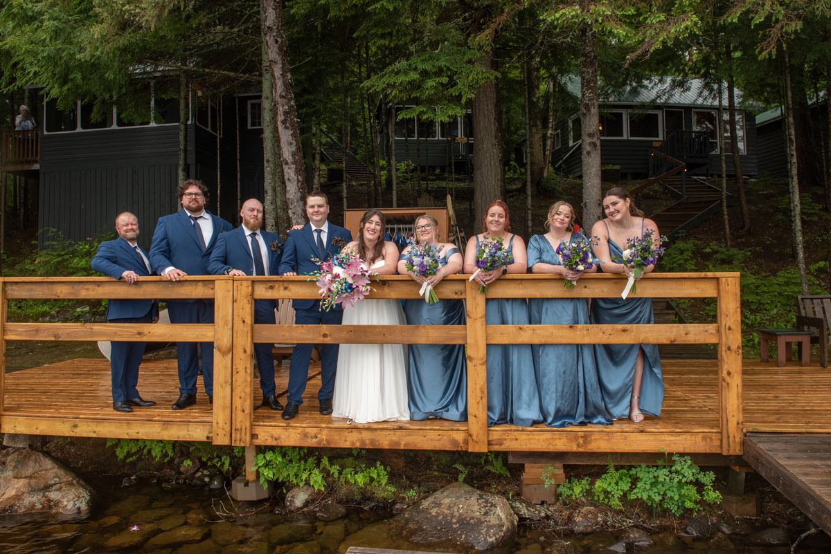 A wedding party poses on a rustic wooden bridge, surrounded by forest and cabins, smiling with bouquets.