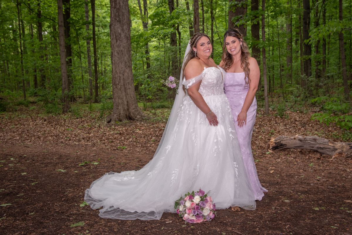 Two brides in forest gowns and veils smile beside pink and white roses on a dirt path surrounded by trees.