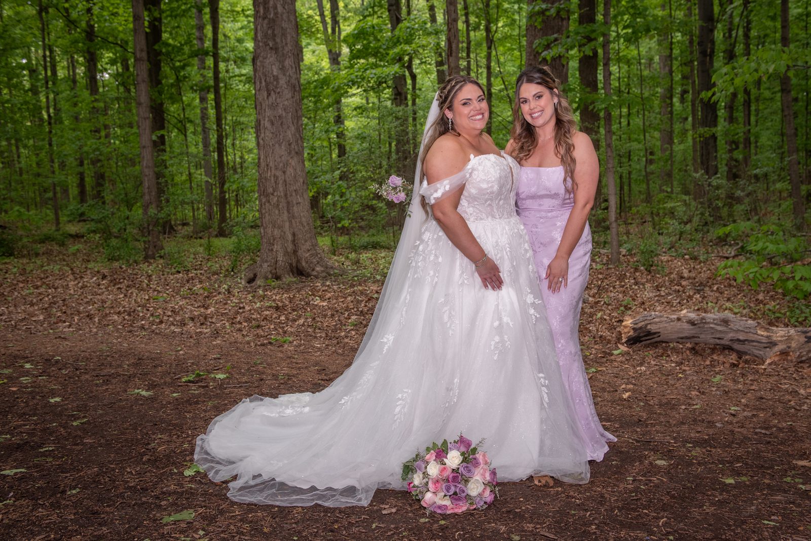 Two brides in forest gowns and veils smile beside pink and white roses on a dirt path surrounded by trees.