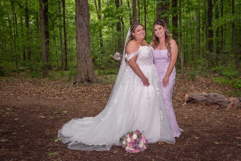 Two brides in forest gowns and veils smile beside pink and white roses on a dirt path surrounded by trees.