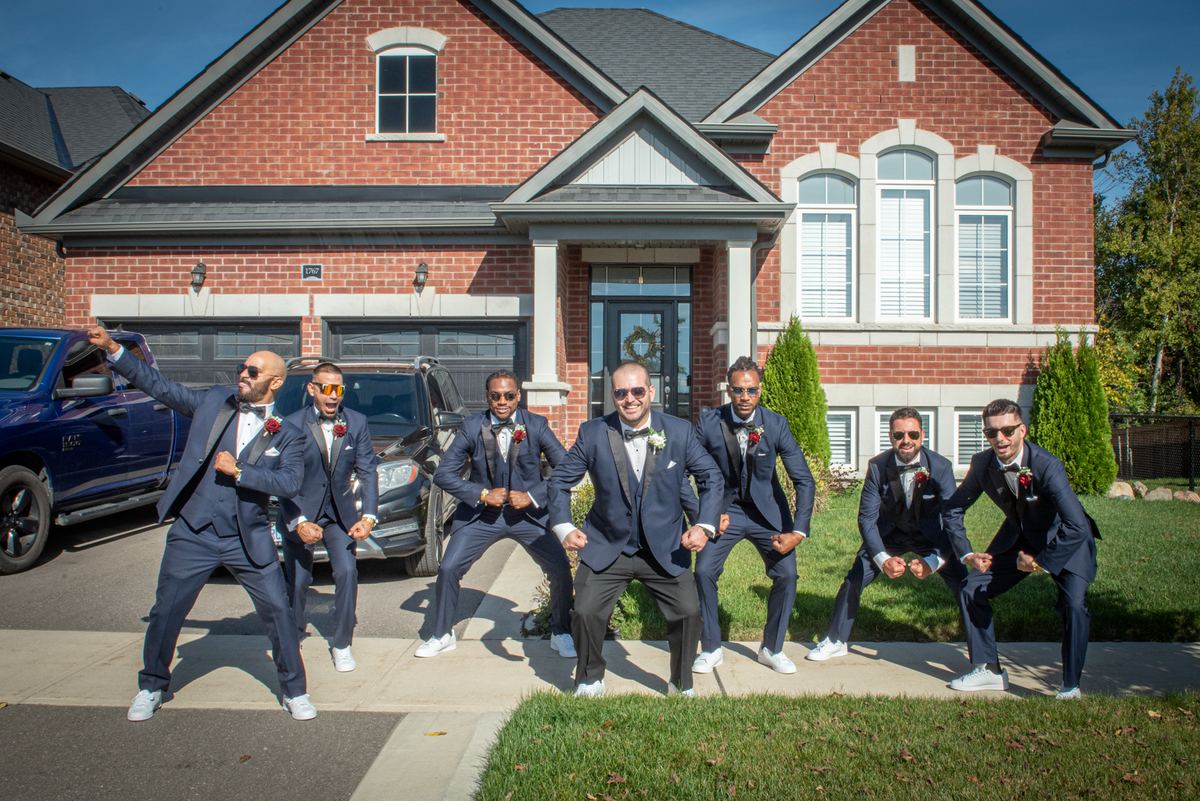 Seven men in navy tuxedos pose playfully on a driveway in front of a red brick house, sunglasses perched atop their heads.