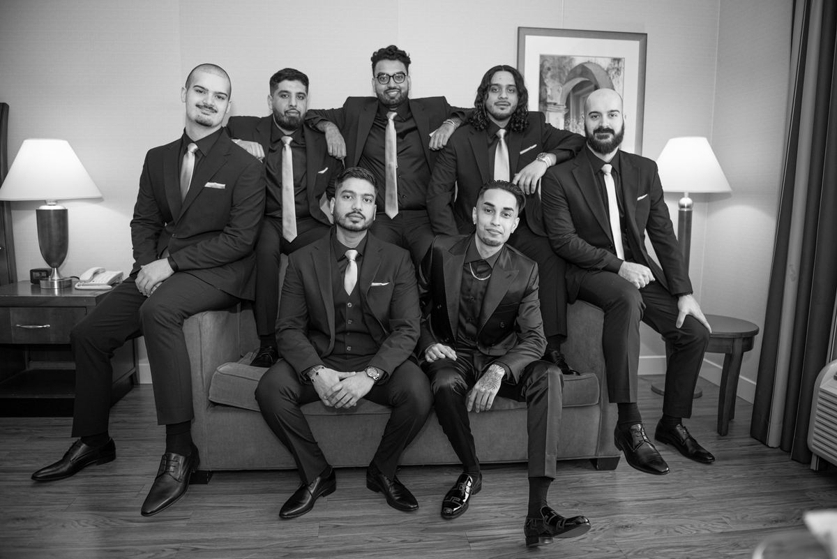 Seven formally dressed men posing together in a hotel room, displaying relaxed camaraderie on and around a sofa.
