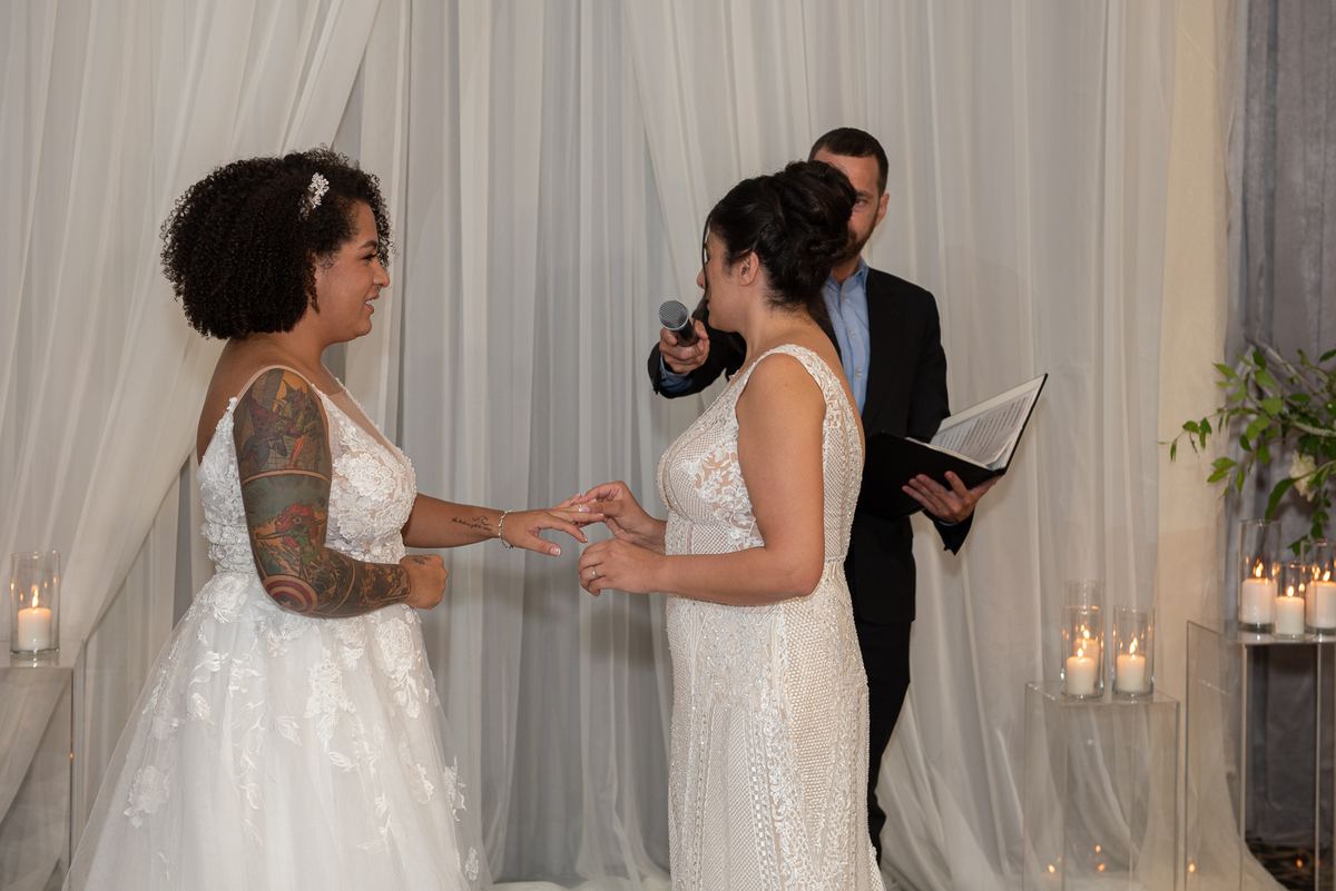 Two brides exchange rings during their ceremony, illuminated by soft candlelight and framed by draped white fabric.