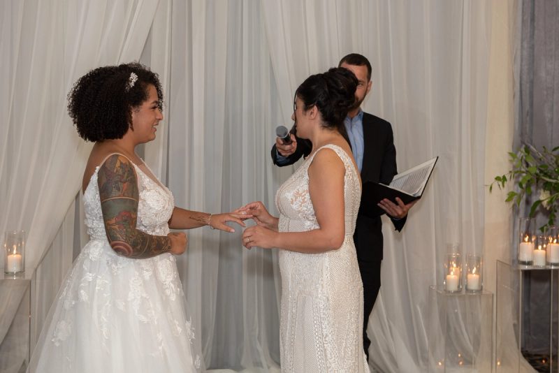 Two brides exchange rings during their ceremony, illuminated by soft candlelight and framed by draped white fabric.