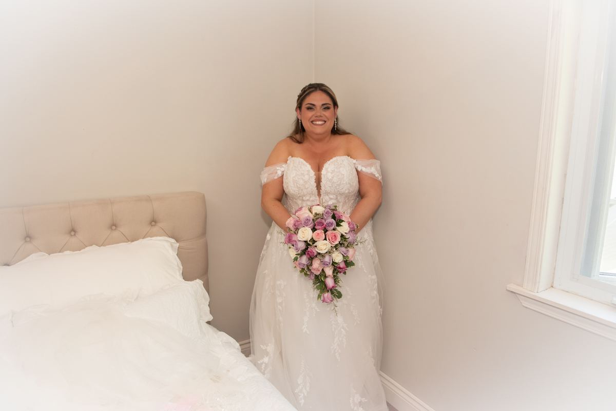 A smiling bride in a bedroom holds pink and white roses, with natural light casting a gentle glow on the scene.bedroom