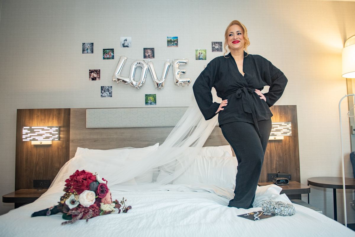 A bride in a black robe and veil stands confidently on a bed, hands on hips, with decorations and wedding items nearby.