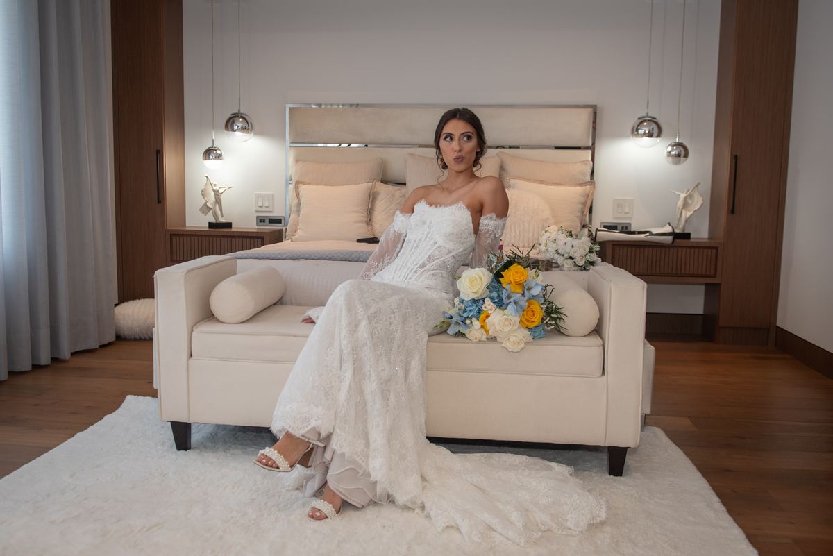 A bride in a white lace gown sits on a cream sofa with a yellow and blue flower bouquet, illuminated by soft lighting.