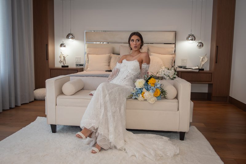 A bride in a white lace gown sits on a cream sofa with a yellow and blue flower bouquet, illuminated by soft lighting.