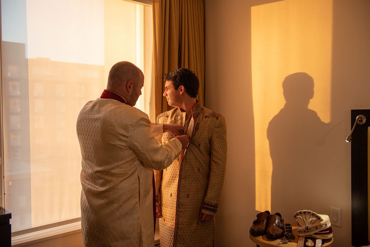A man helps another into a traditional embroidered sherwani in a warmly lit room, sunlight casting long shadows on the wall.