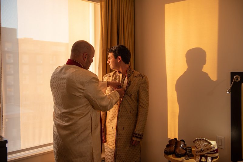 A man helps another into a traditional embroidered sherwani in a warmly lit room, sunlight casting long shadows on the wall.