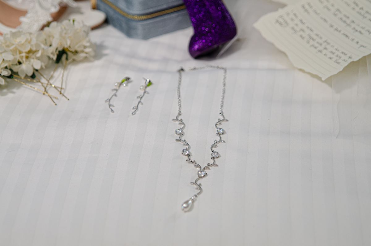A silver necklace and earrings lie on white fabric with flowers and a note, creating an intimate gift scene.