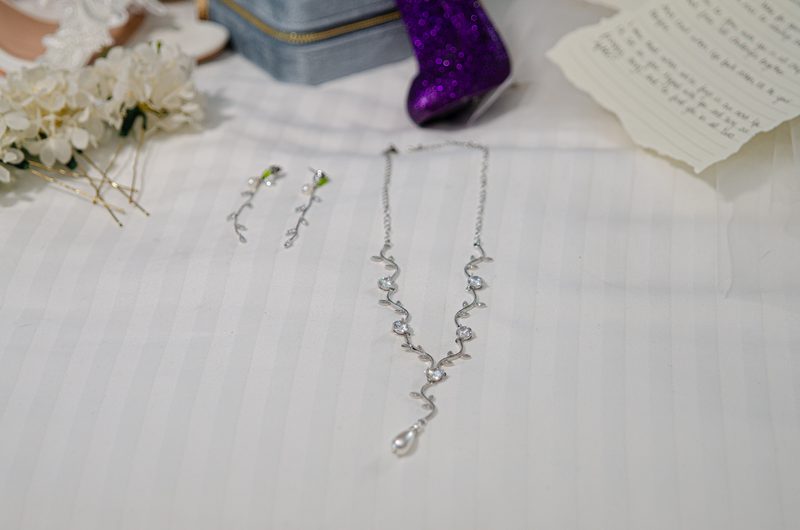 A silver necklace and earrings lie on white fabric with flowers and a note, creating an intimate gift scene.
