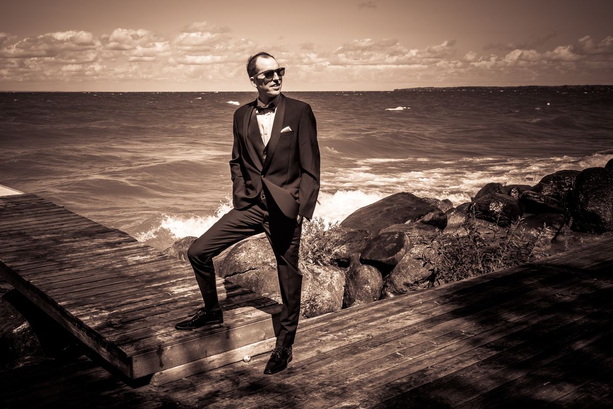 A man in a tuxedo and sunglasses leans on a wooden dock beside rocks, with waves crashing behind him.