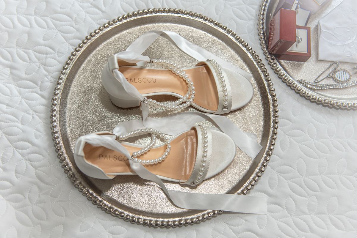 Elegant pearl-embellished white wedding shoes resting on a silver tray atop quilted fabric, illuminated by soft light.