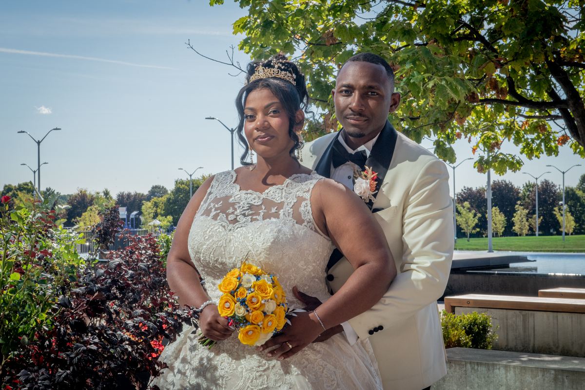 A bride in a lace gown and tiara with a bouquet stands beside her groom in a cream tuxedo, surrounded by greenery.