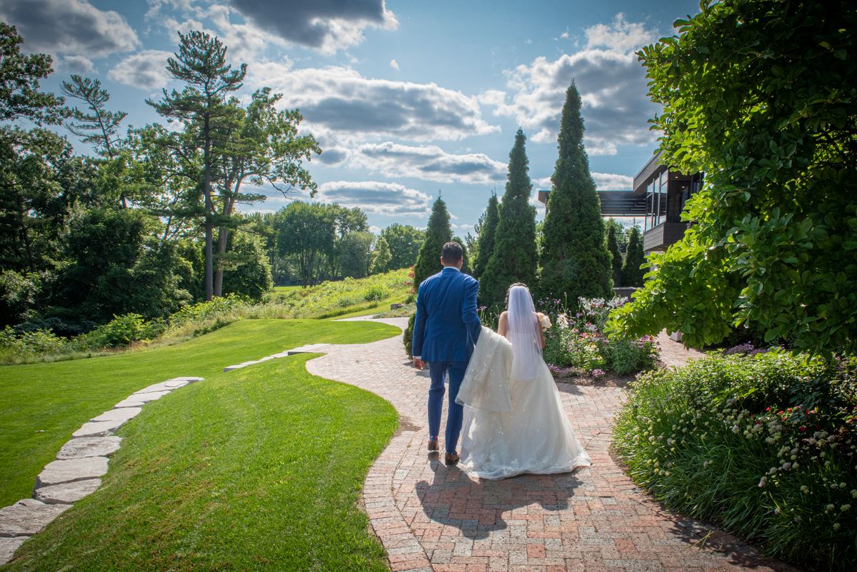 A bride and groom walk down a brick path in a sunlit garden toward a modern building surrounded by evergreens.