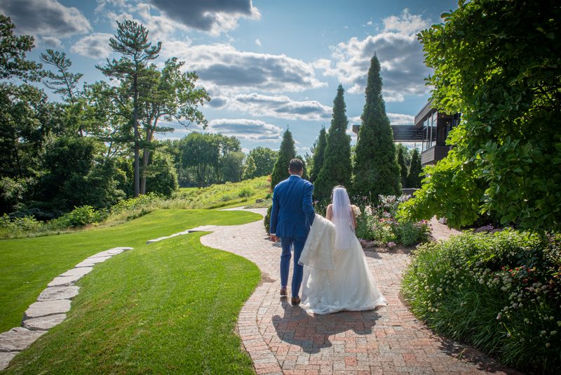 A bride and groom walk down a brick path in a sunlit garden toward a modern building surrounded by evergreens.