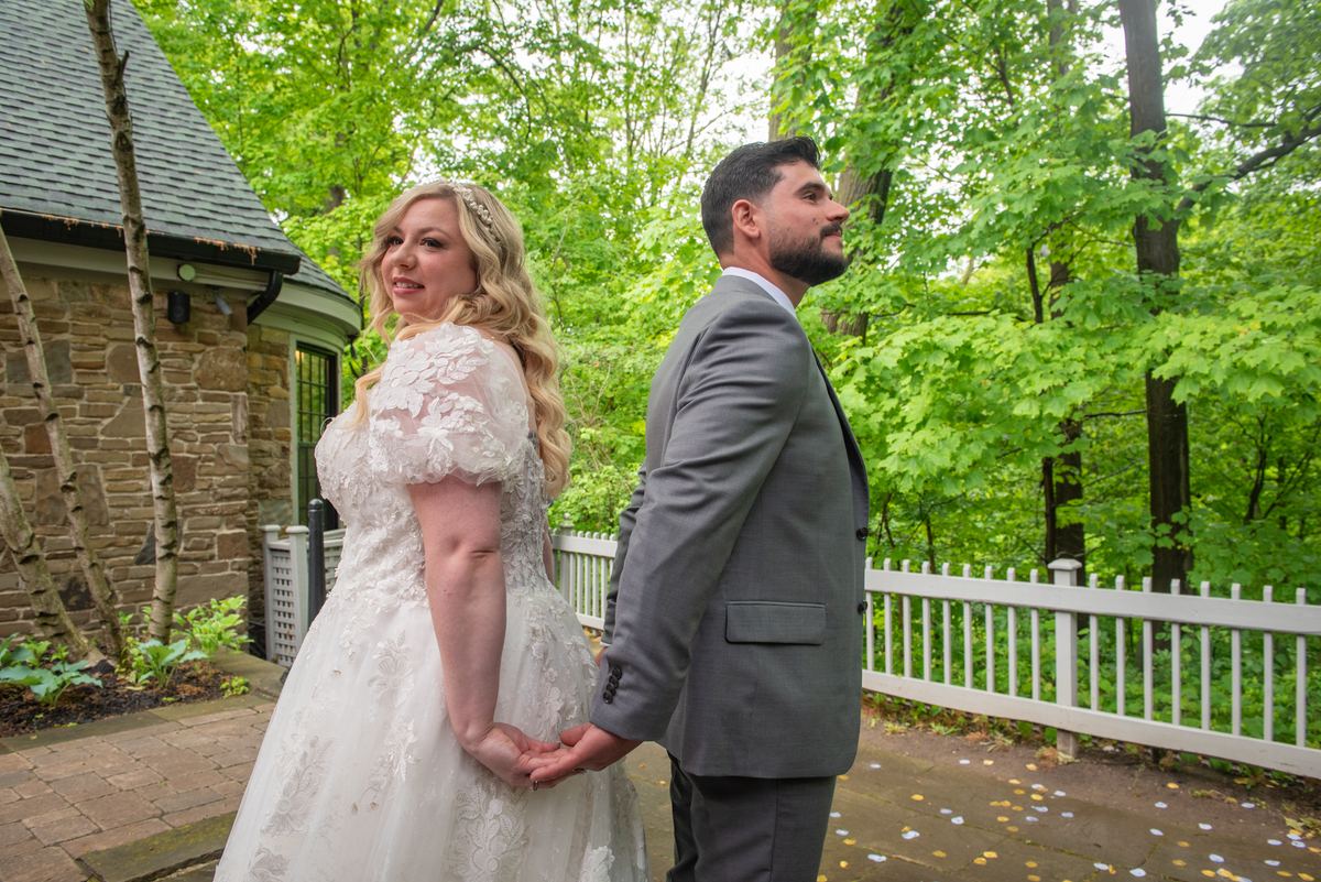 A bride and groom stand hand-in-hand near a stone building, framed by lush trees and white picket fence.