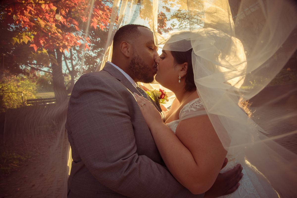 A newlywed couple kisses outdoors, framed by the bride's veil and golden sunlight, with autumn leaves in the background.