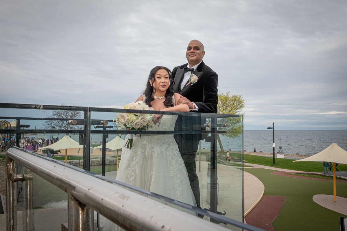 Bride and groom stand on a glass-railled overlook, gazing at the camera as people gather under umbrellas near the waterfront.