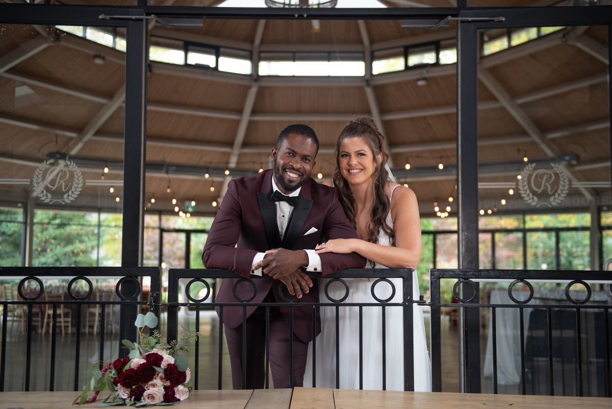 Bride in white and groom in burgundy tuxedo smile beside black railing, illuminated by natural light.