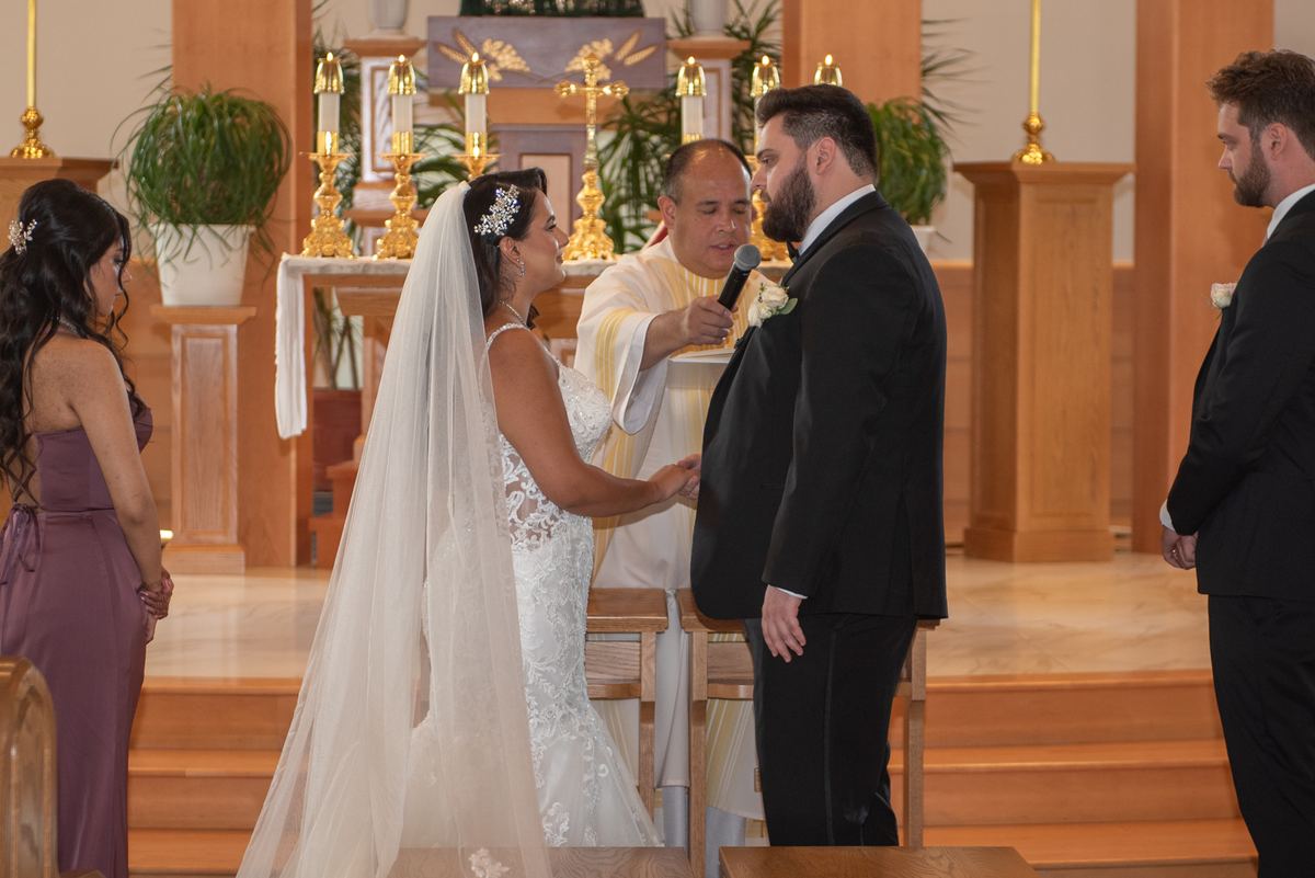 A bride and groom stand at an altar, holding hands, as a priest speaks, with candles and a bridesmaid in purple nearby.