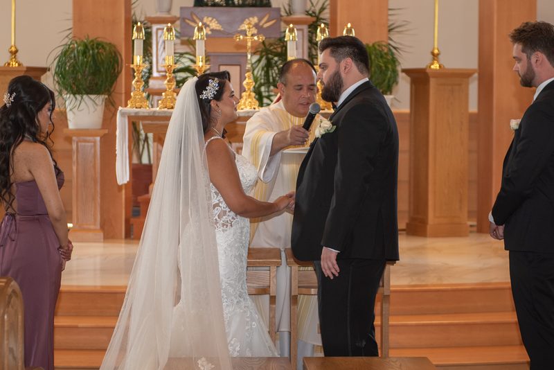 A bride and groom stand at an altar, holding hands, as a priest speaks, with candles and a bridesmaid in purple nearby.