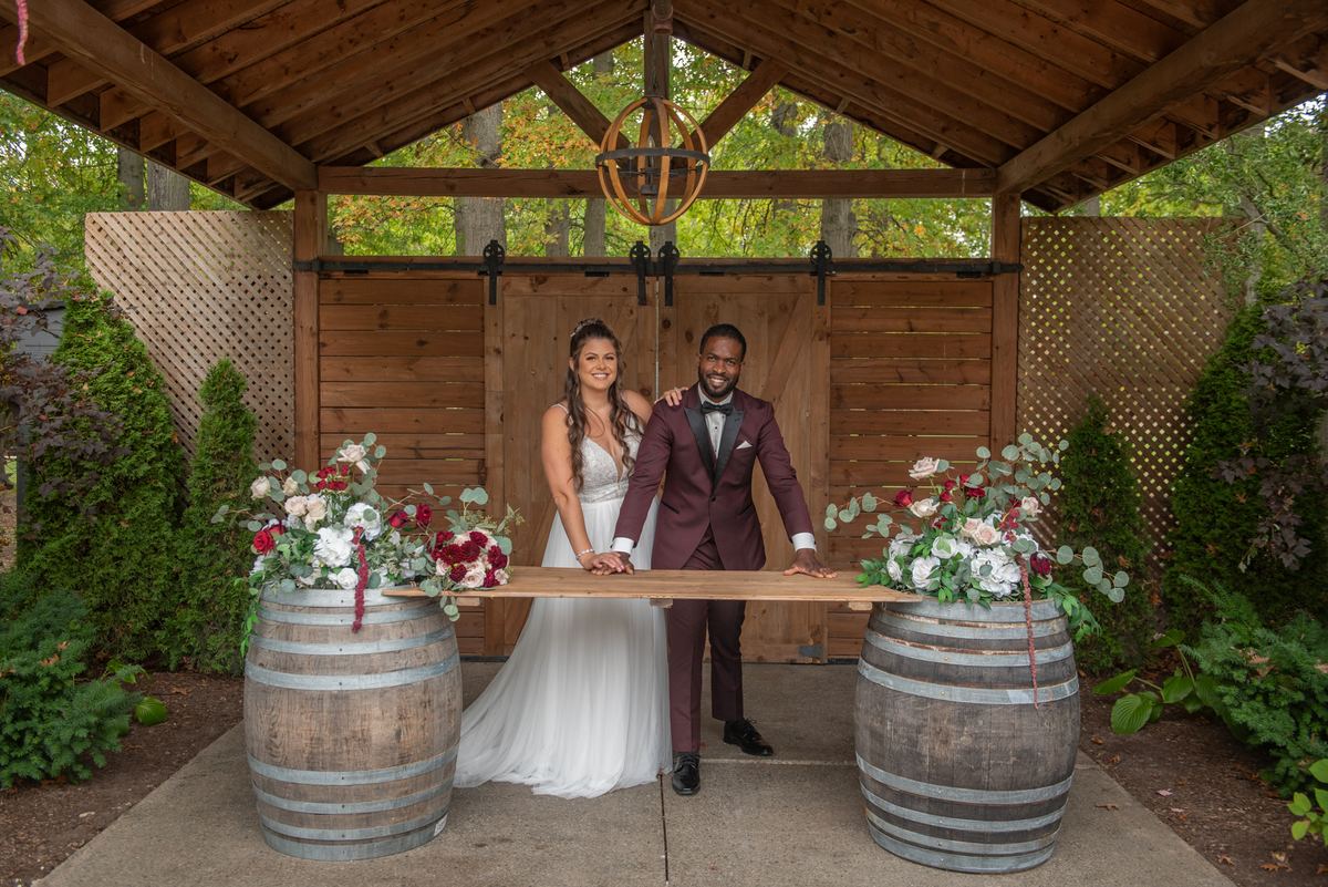 A smiling bride and groom hold a wooden plank over wine barrels under a pavilion, surrounded by autumn foliage.