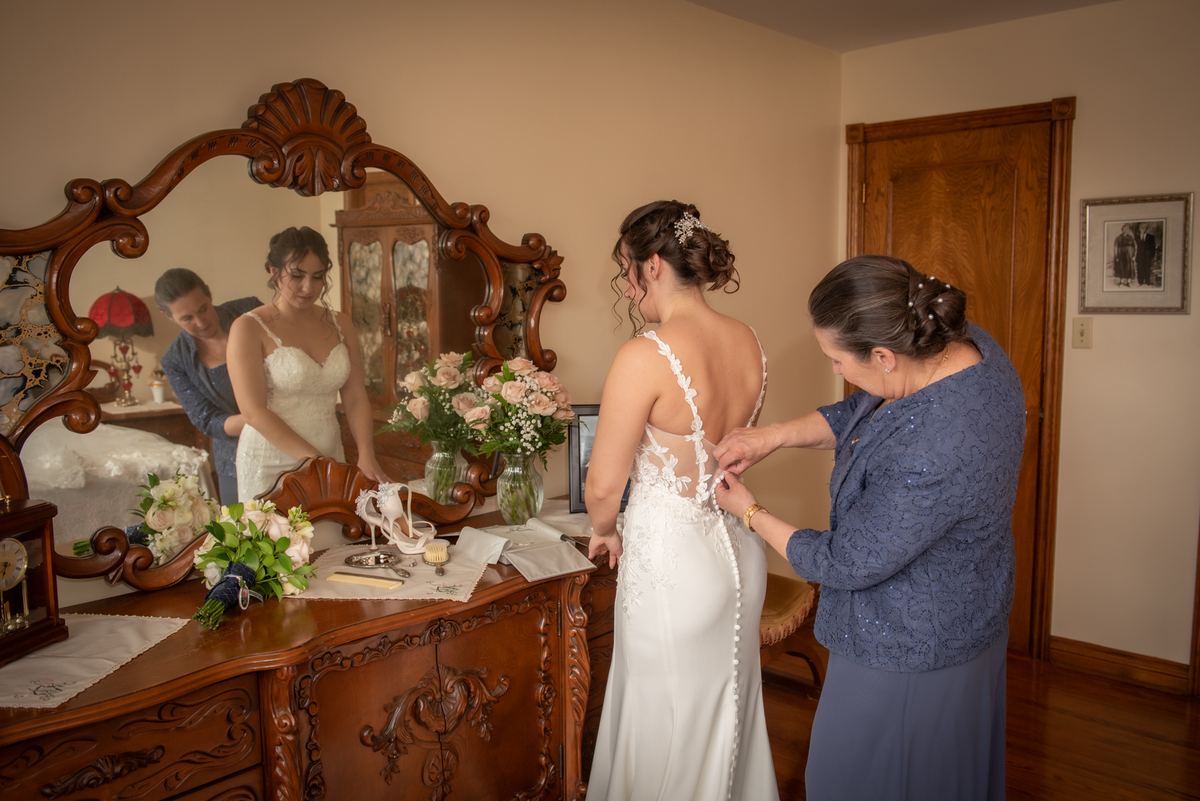 A bride stands as her mother fastens her wedding gown in a warmly lit bedroom with floral arrangements and wooden furniture.