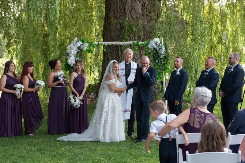 Bride and groom exchange vows under a willow tree, surrounded by smiling family members and bridesmaids in purple gowns.