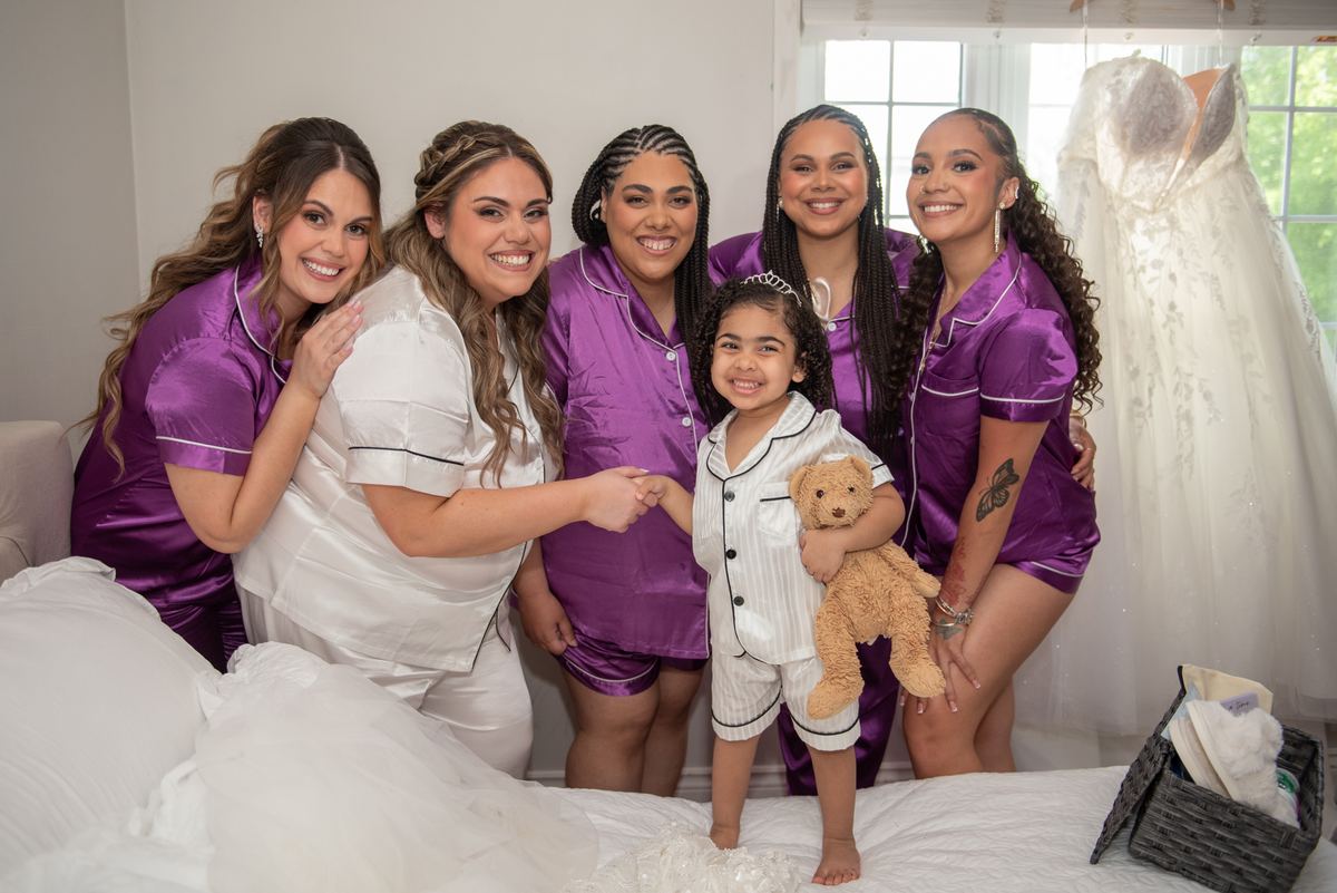 Bride and bridesmaids in pajamas smile with a young girl holding a teddy bear beside a wedding gown.