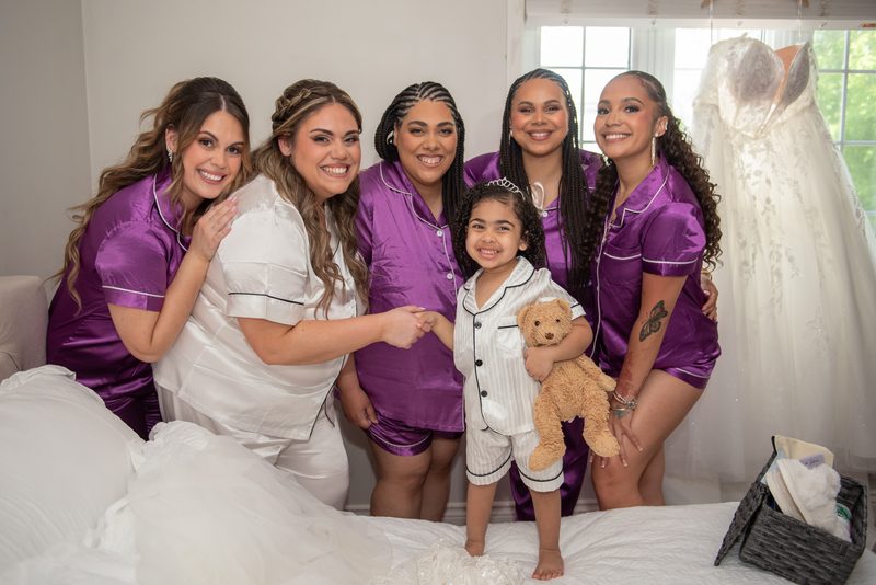 Bride and bridesmaids in pajamas smile with a young girl holding a teddy bear beside a wedding gown.